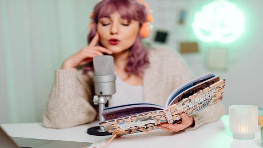 Audiobook voice over jobs. Image of a young woman recording a children's story audiobook in front of a microphone. This image is used to illustrate the article "Audiobook Voice Over Jobs: An Emerging Opportunity." Pexels license: https://www.pexels.com/photo/woman-in-brown-sweater-holding-a-book-6919969/