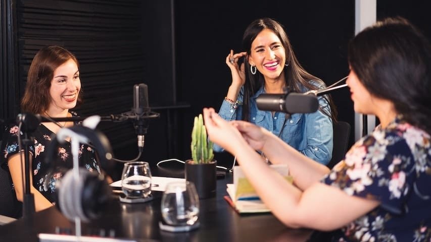 Types of Podcasts. Image of three women around a table with microphones recording a podcast. This image is used to illustrate the article "Exploring the 3 Types of Podcasts". Pexels license: https://www.pexels.com/photo/women-sitting-at-the-table-7586484/
