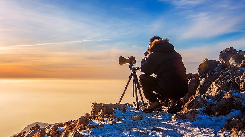 How to Do Voice Over for Documentary. Photo of professional photographer takes photos with camera on tripod on rocky peak at sunset. Image licence from FreePik: https://www.freepik.com/free-photo/professional-photographer-takes-photos-with-camera-tripod-rocky-peak-sunset-dark-tone_11599632.htm This image is being used to illustrate the article "How to Do Voice Over for Documentary Films."